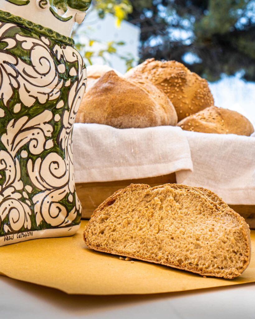 Fetta di pane integrale a lievito madre con mollica alveolata, davanti a un cesto di pagnotte e a una ceramica decorata